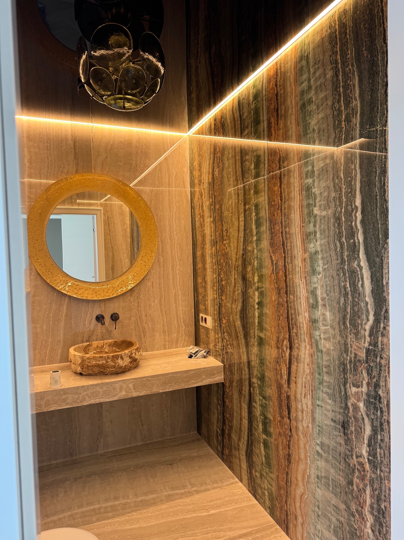 Oryginalna łazienka z kamienną umywalką i okrągłym lustrem podświetlonym ledami. Ściany wyłożone płytami z wzorem imitującym naturalny kamień.
