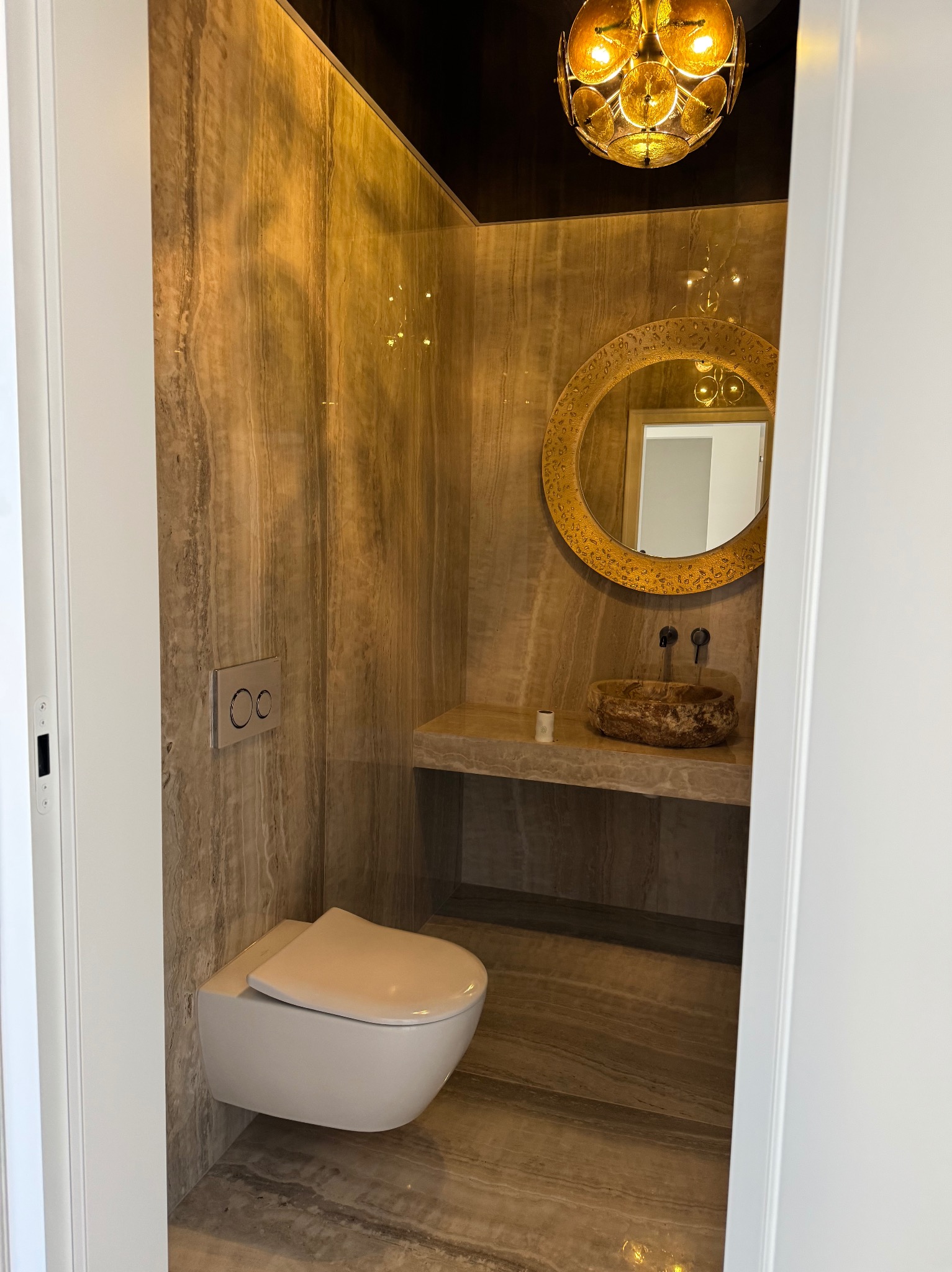 Wykończona łazienka z beżowymi płytkami imitującymi marmur, wiszącą toaletą, kamienną umywalką i okrągłym lustrem w złotej ramie, widok zza białych drzwi.