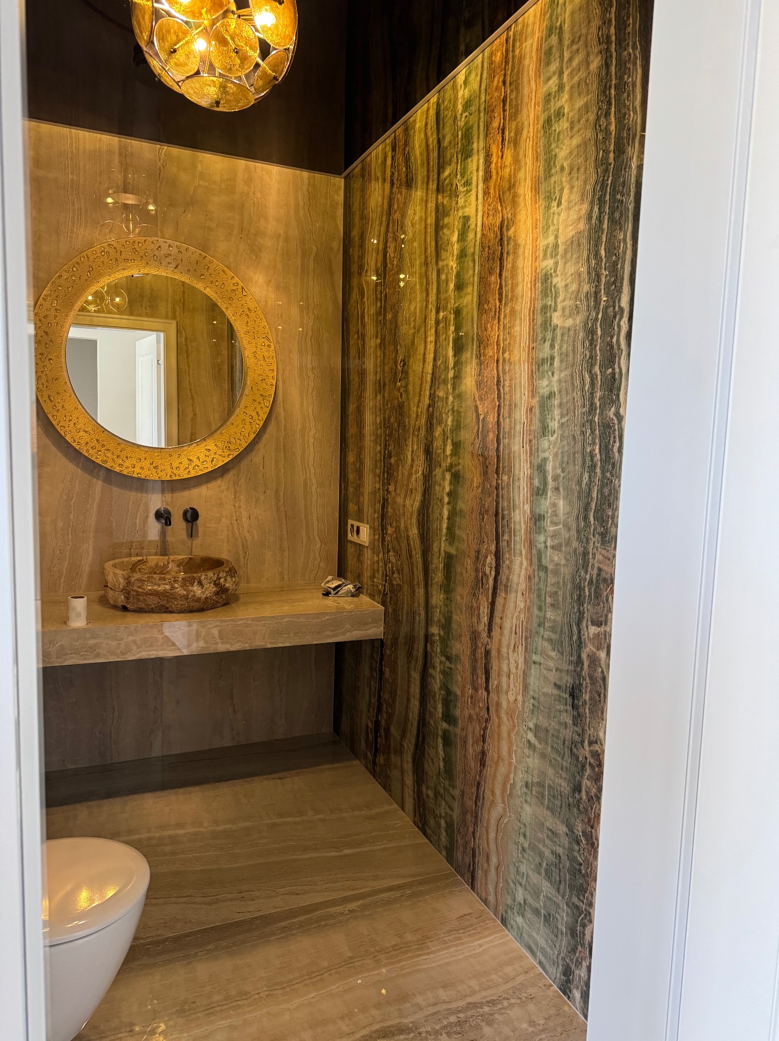 Oryginalna łazienka z umywalką z kamienia, okrągłym lustrem w złotej ramie i ścianą z dekoracyjnych paneli imitujących drewno w odcieniach brązu i zieleni.