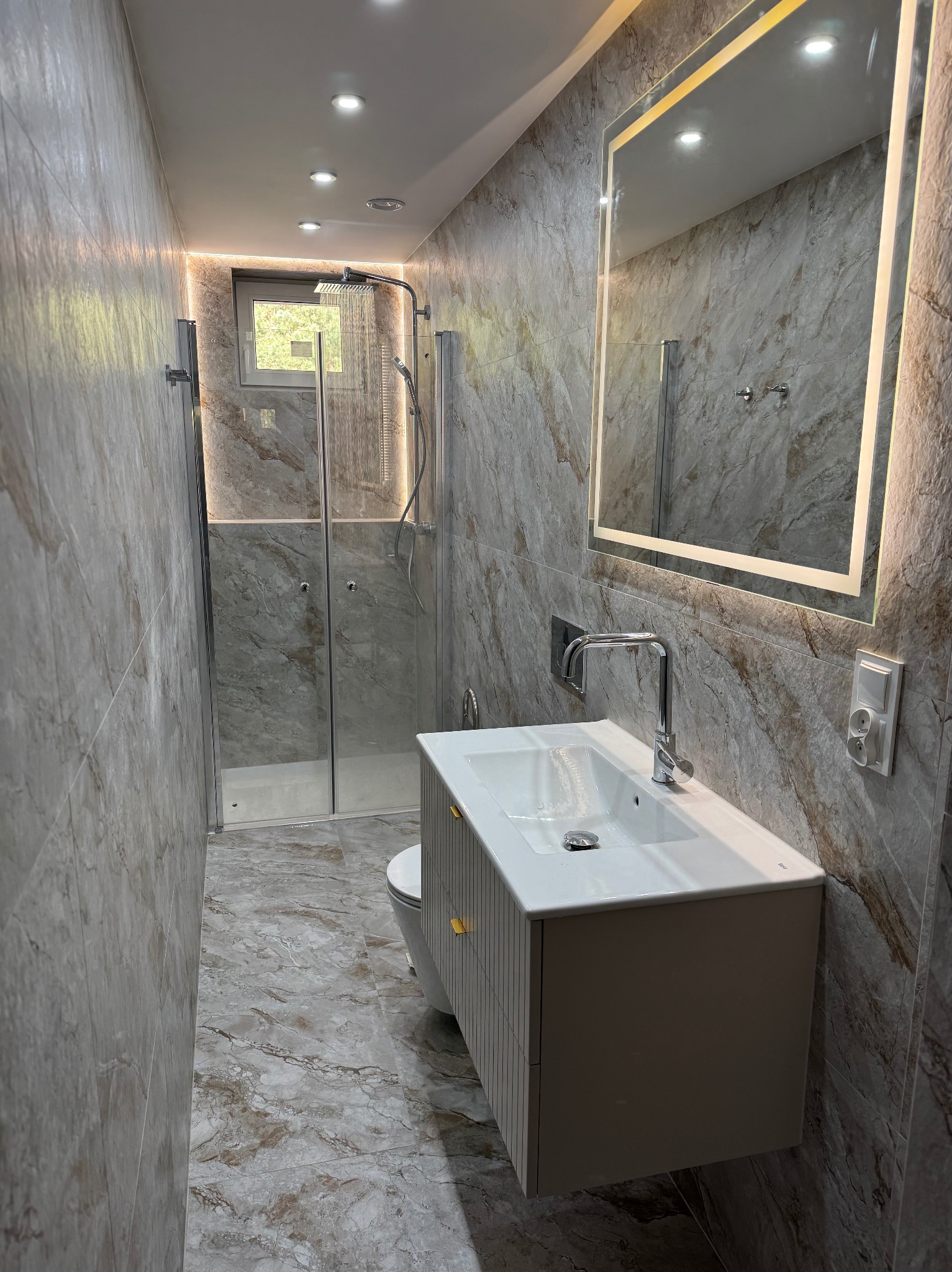 Wykończona łazienka z kabiną prysznicową, umywalką z szafką i podświetlanym lustrem. Aranżacja w odcieniach szarości z marmurkową fakturą płytek. Nowoczesny design i oświetlenie.