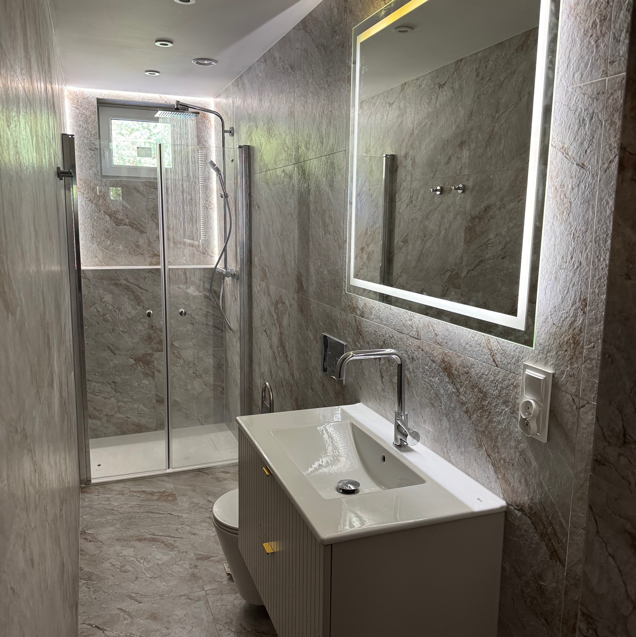 Wykończona łazienka z kabiną prysznicową, umywalką z szafką i podświetlanym lustrem. Ściany i podłoga wyłożone płytkami imitującymi marmur.