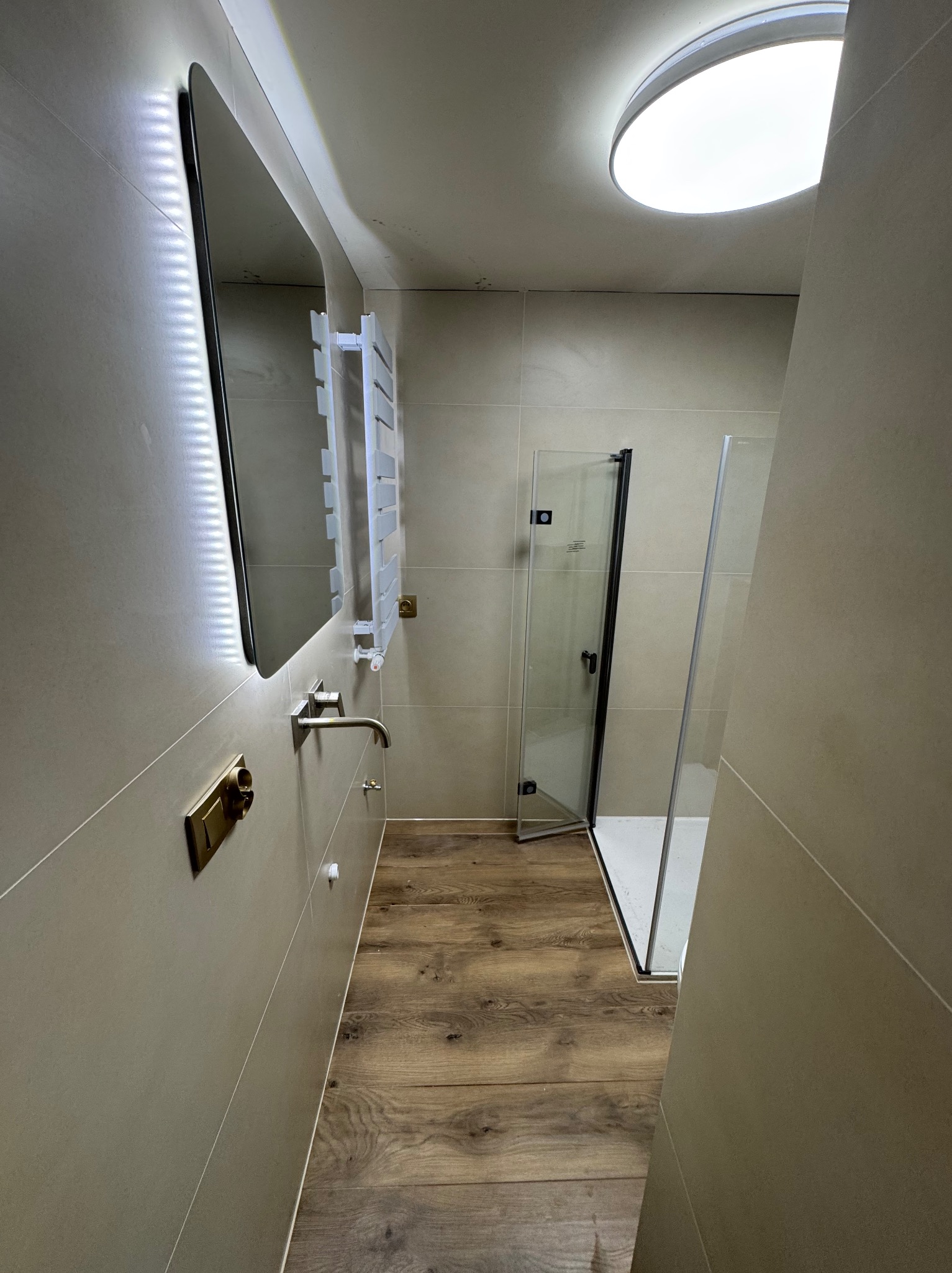 Nowoczesna łazienka po remoncie: beżowe płytki, drewnopodobna podłoga, szklana kabina prysznicowa i podświetlane lustro. Minimalistyczny design z dbałością o detale.