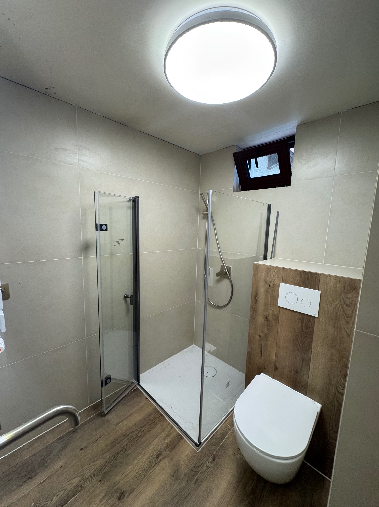Nowoczesna łazienka z kabiną prysznicową typu walk-in, wiszącą toaletą i drewnopodobnymi panelami. Małe okno w górnej części ściany. Całość oświetlona okrągłą lampą sufitową.