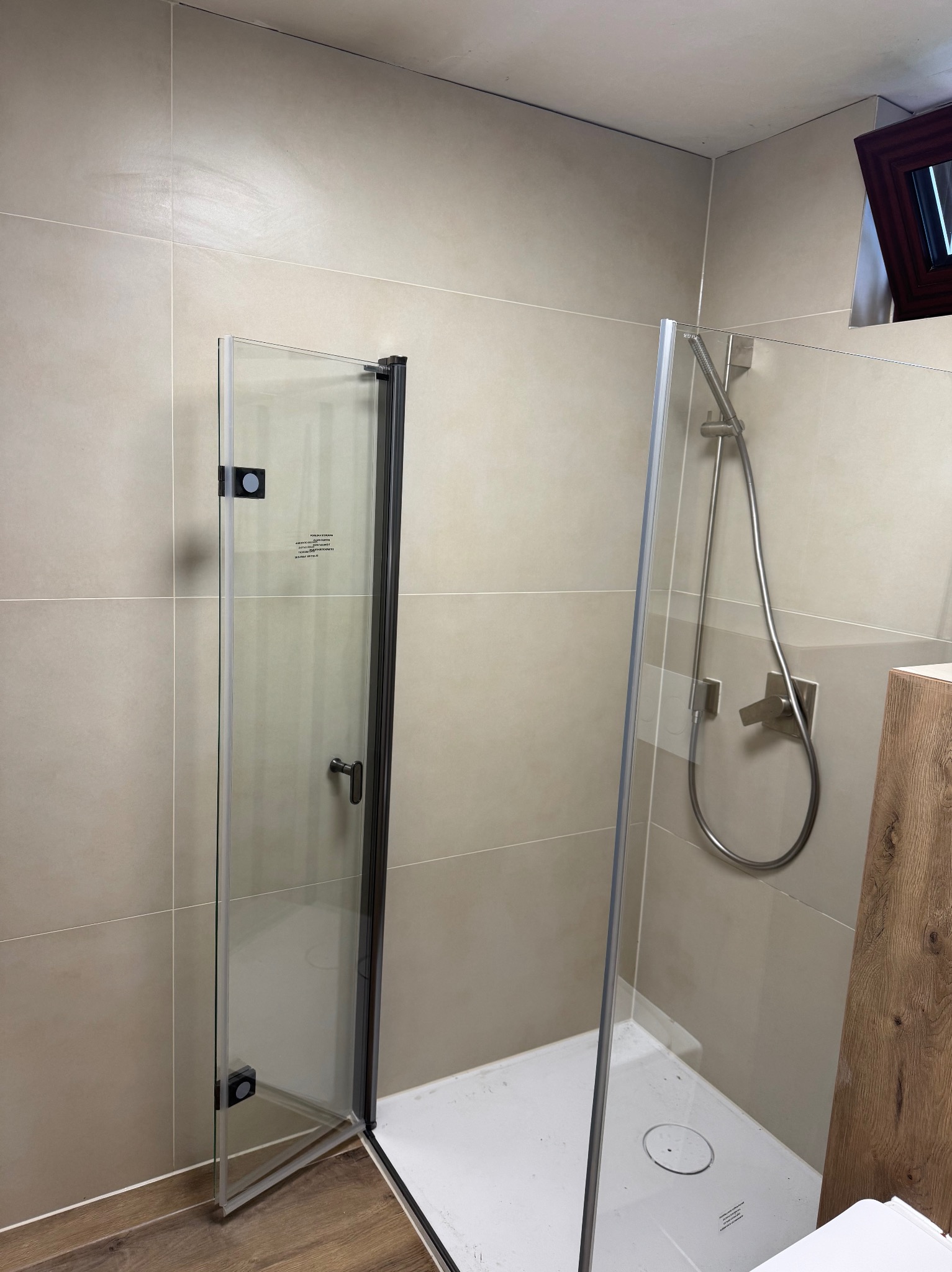 Beżowa łazienka z kabiną prysznicową typu walk-in i częściowo otwartymi drzwiami. Widoczna armatura i płytki. Minimalistyczny design.