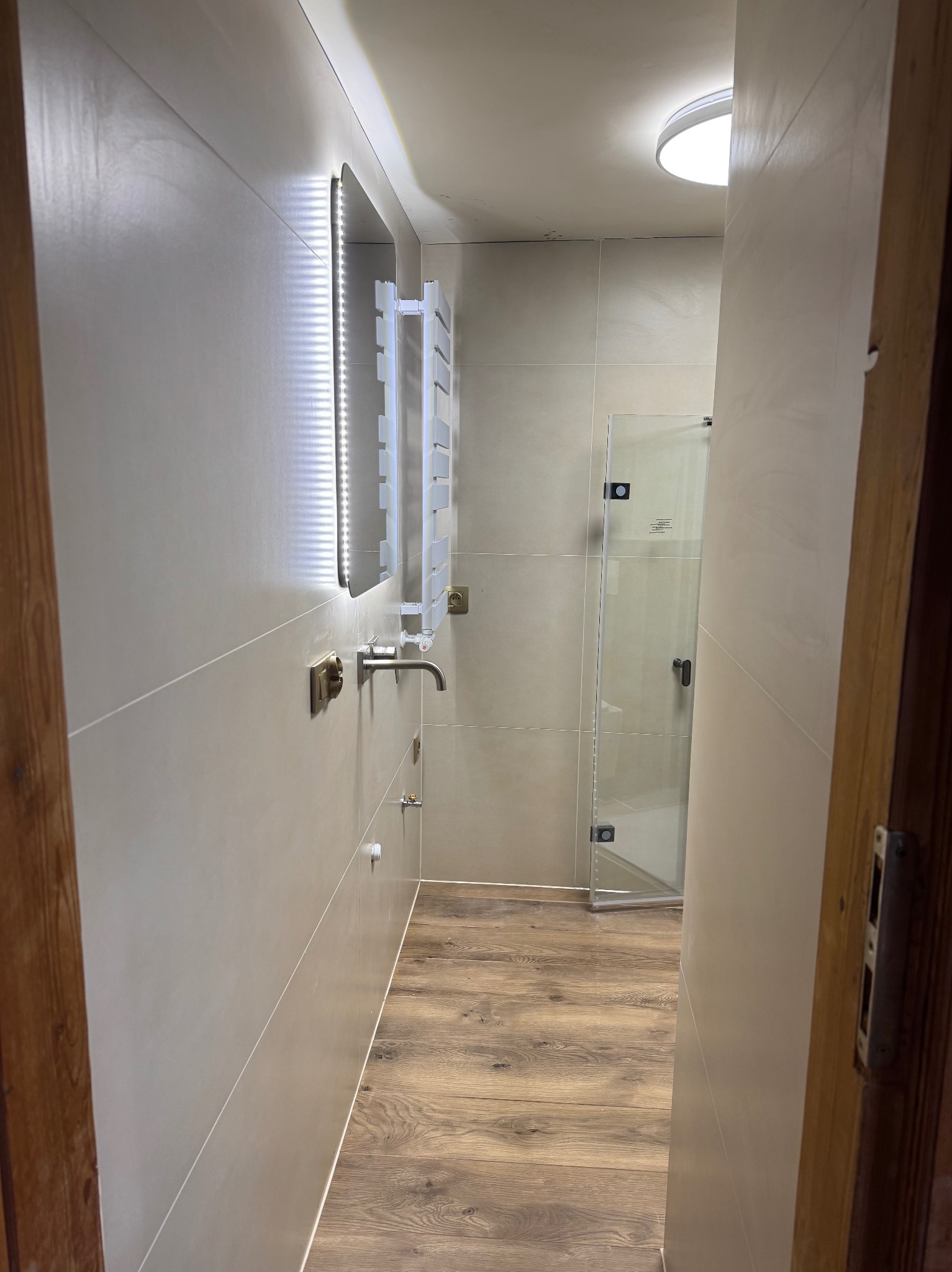 Jasna łazienka po remoncie z podświetlanym lustrem, grzejnikiem drabinkowym i kabiną prysznicową. Widoczna drewnopodobna podłoga i płytki w odcieniach szarości.