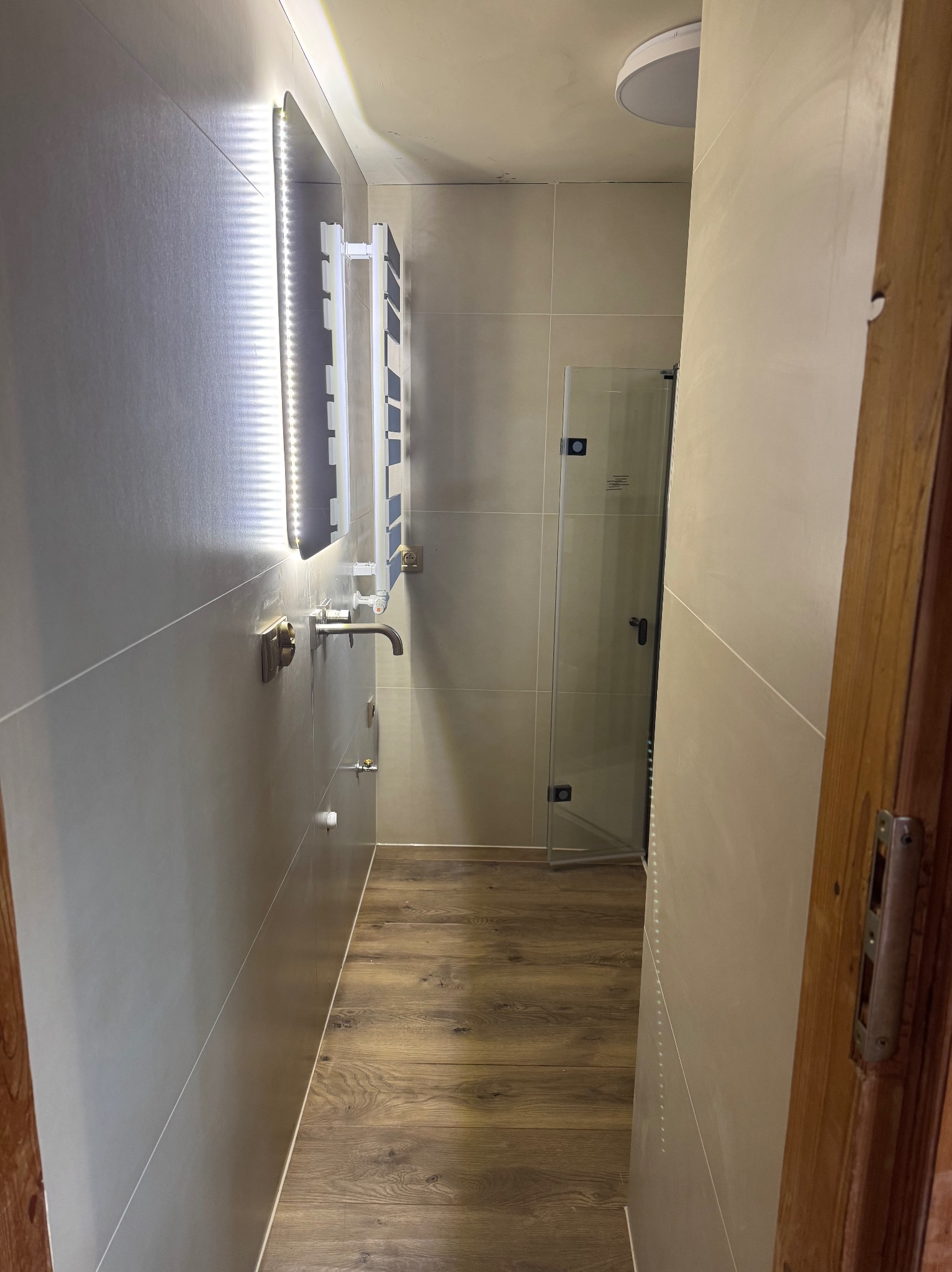 Wąska, wykafelkowana łazienka z podświetlanym lustrem i drewnopodobną podłogą, widok zza uchylonych drewnianych drzwi. Kabina prysznicowa ze szkła.