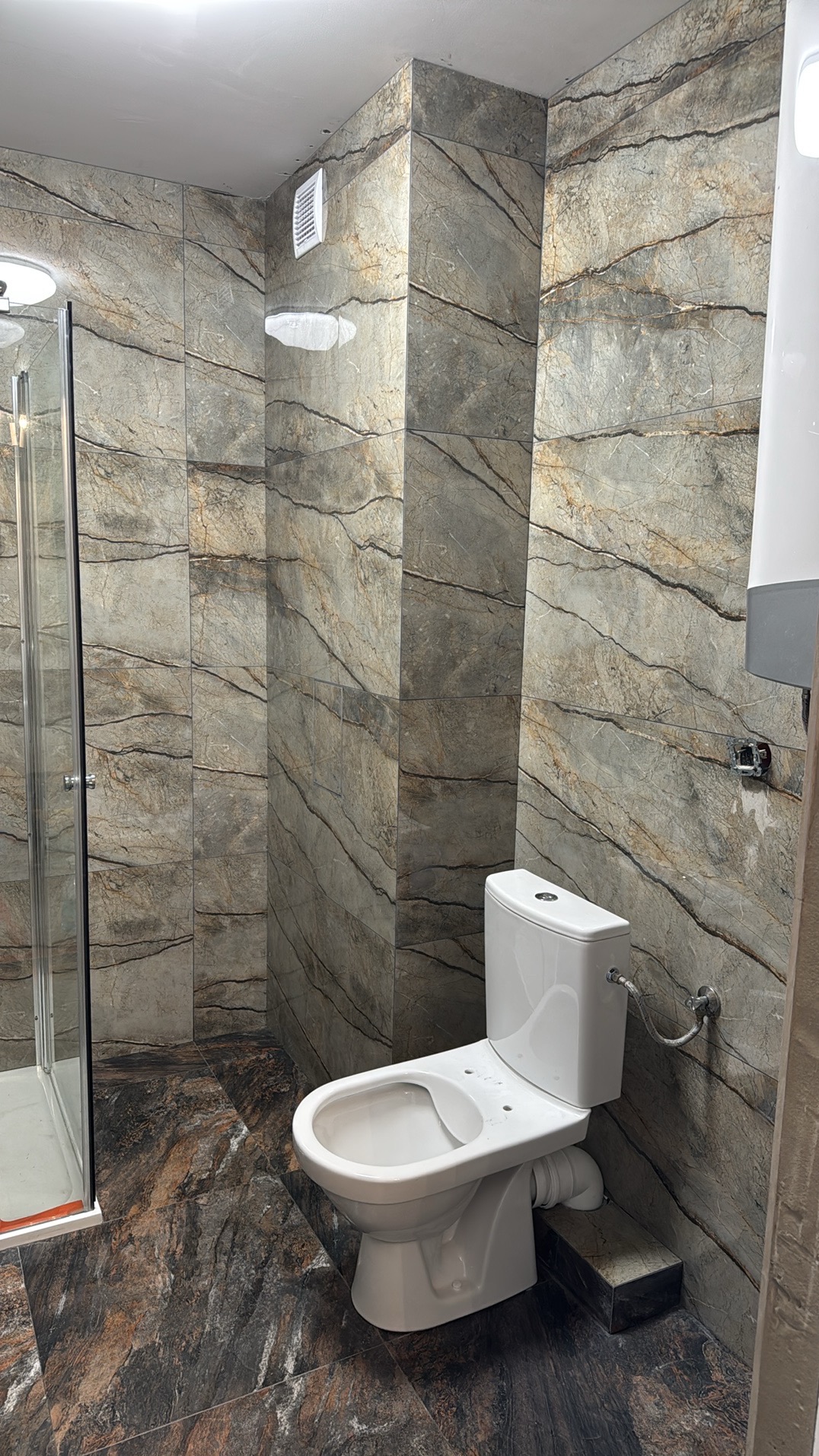Wykończona łazienka z marmurowymi płytkami, kabiną prysznicową i podwieszaną toaletą. Widoczne detale instalacji i wentylacja.