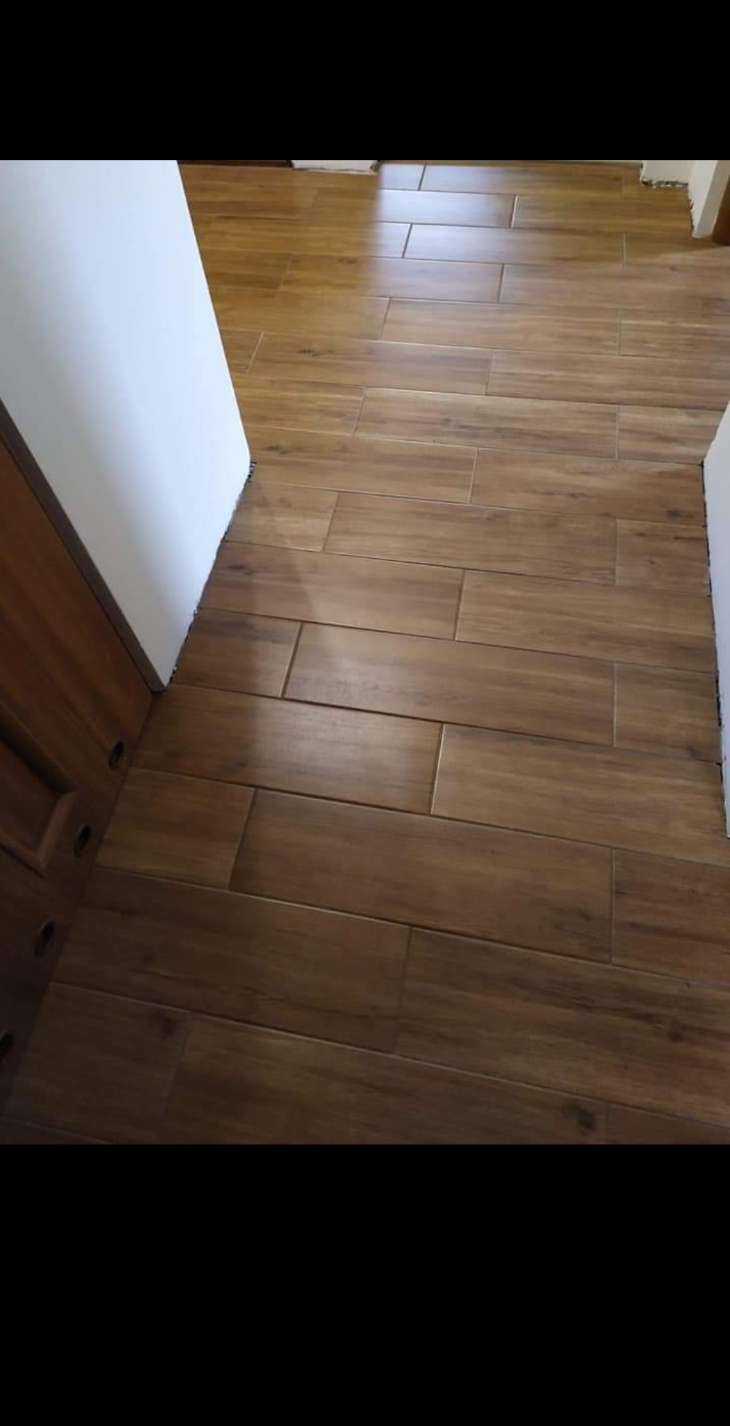 Ułożona podłoga z podłużnych, brązowych płytek imitujących drewno, widoczne fragmenty białych ścian i brązowych mebli.