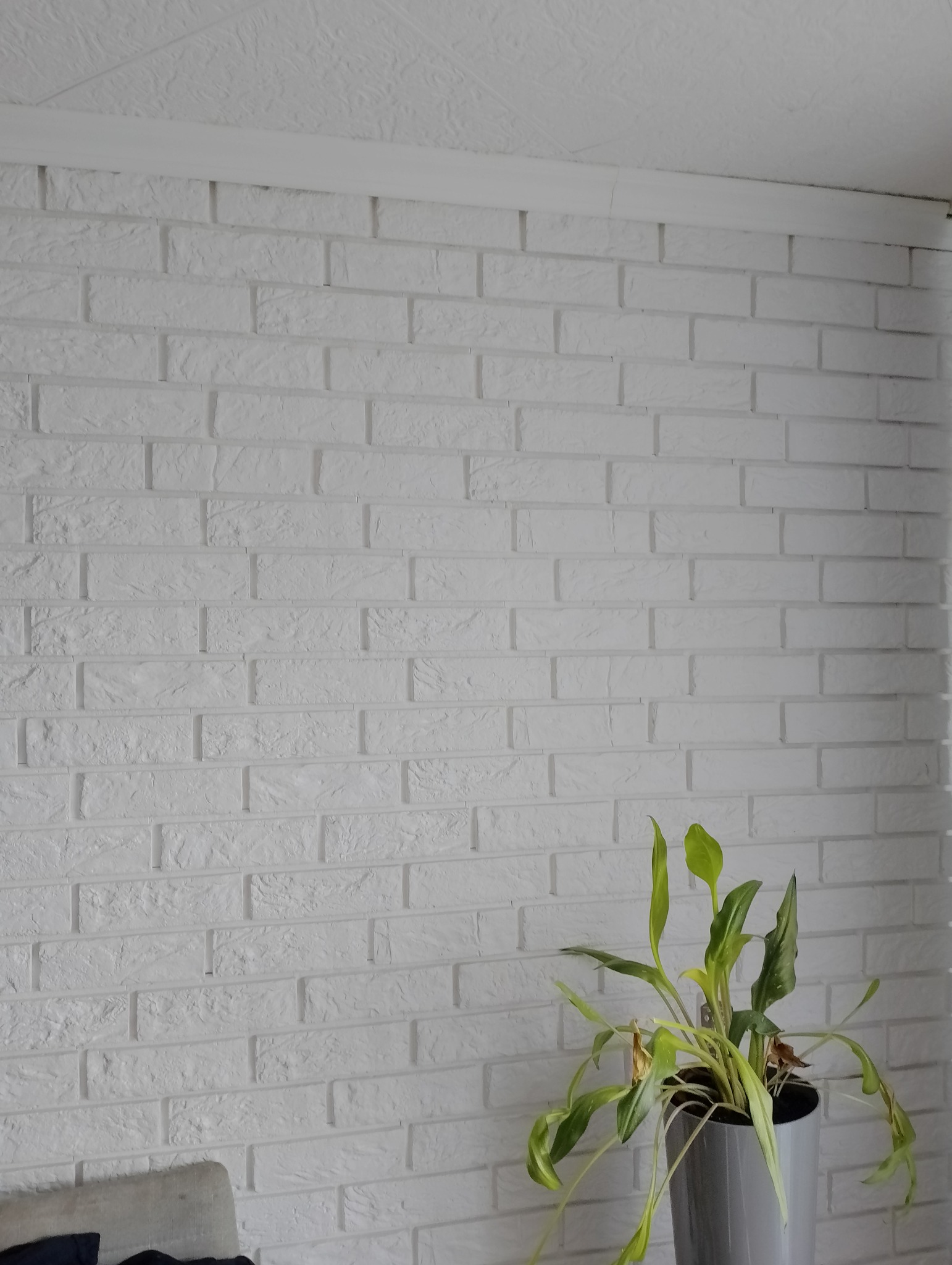 Ściana z białej cegły dekoracyjnej z widocznym fragmentem sufitu i rośliną doniczkową w szarej osłonce.