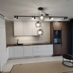 Nowoczesna kuchnia w Krakowie z białymi szafkami, drewnianymi akcentami i designerską lampą wiszącą z trzema szklanymi kloszami. Widoczne elementy wykończenia wnętrz, takie jak ciemne zasłony...