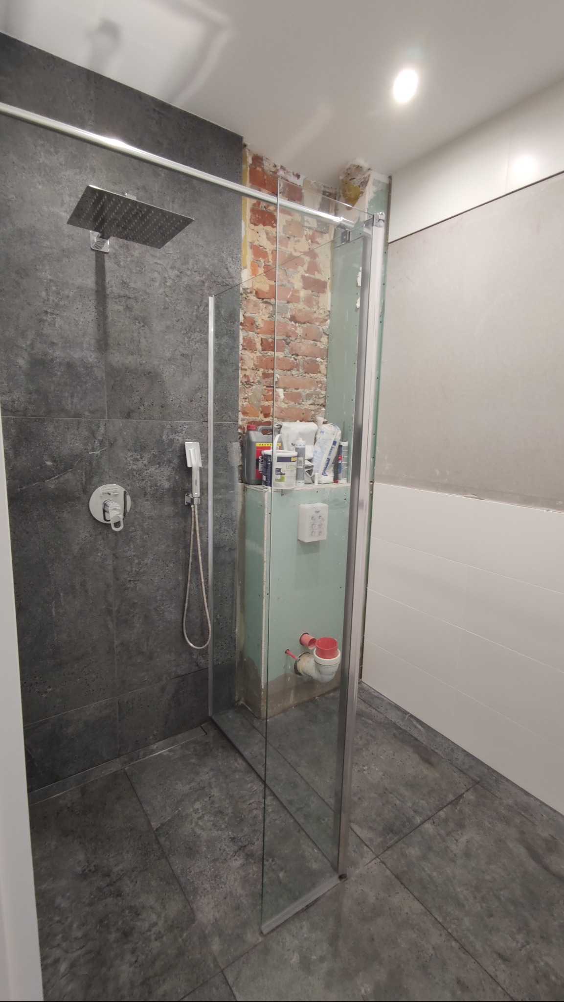 Niedokończona łazienka z szarą, teksturowaną kabiną prysznicową typu walk-in, odsłoniętym murem z cegły i instalacją wodno-kanalizacyjną w trakcie montażu.