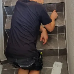 Pracownik w czarnej koszulce fuguje szare płytki na schodach wewnętrznych, widoczne narzędzia: silikon i skrobak do fug.