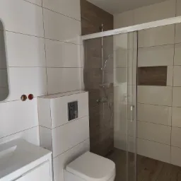 Nowoczesna łazienka z kabiną prysznicową ze szklanymi drzwiami przesuwnymi, toaletą podwieszaną, umywalką z szafką i białymi płytkami na ścianach z brązowymi fugami oraz imitacją drewna w strefie...