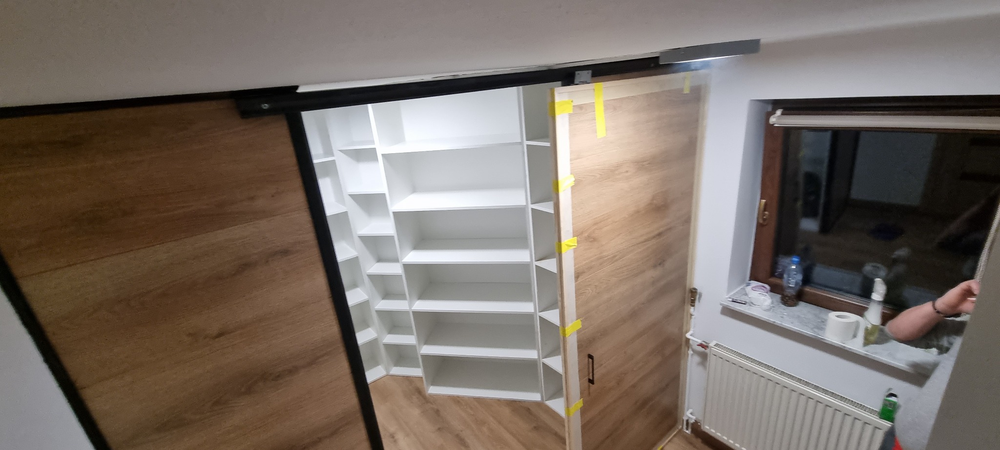 Garderoba na poddaszu z przesuwnymi drzwiami z okleiną drewnopodobną, białe półki, widoczne taśmy zabezpieczające na drzwiach, kaloryfer pod oknem.