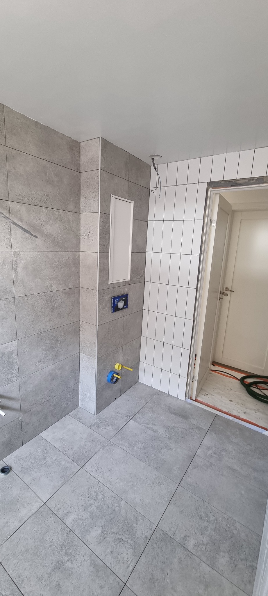 Szara łazienka w trakcie remontu, widoczne płytki na ścianach i podłodze, instalacja podtynkowa WC, wystające rury wodne, brak białego montażu.