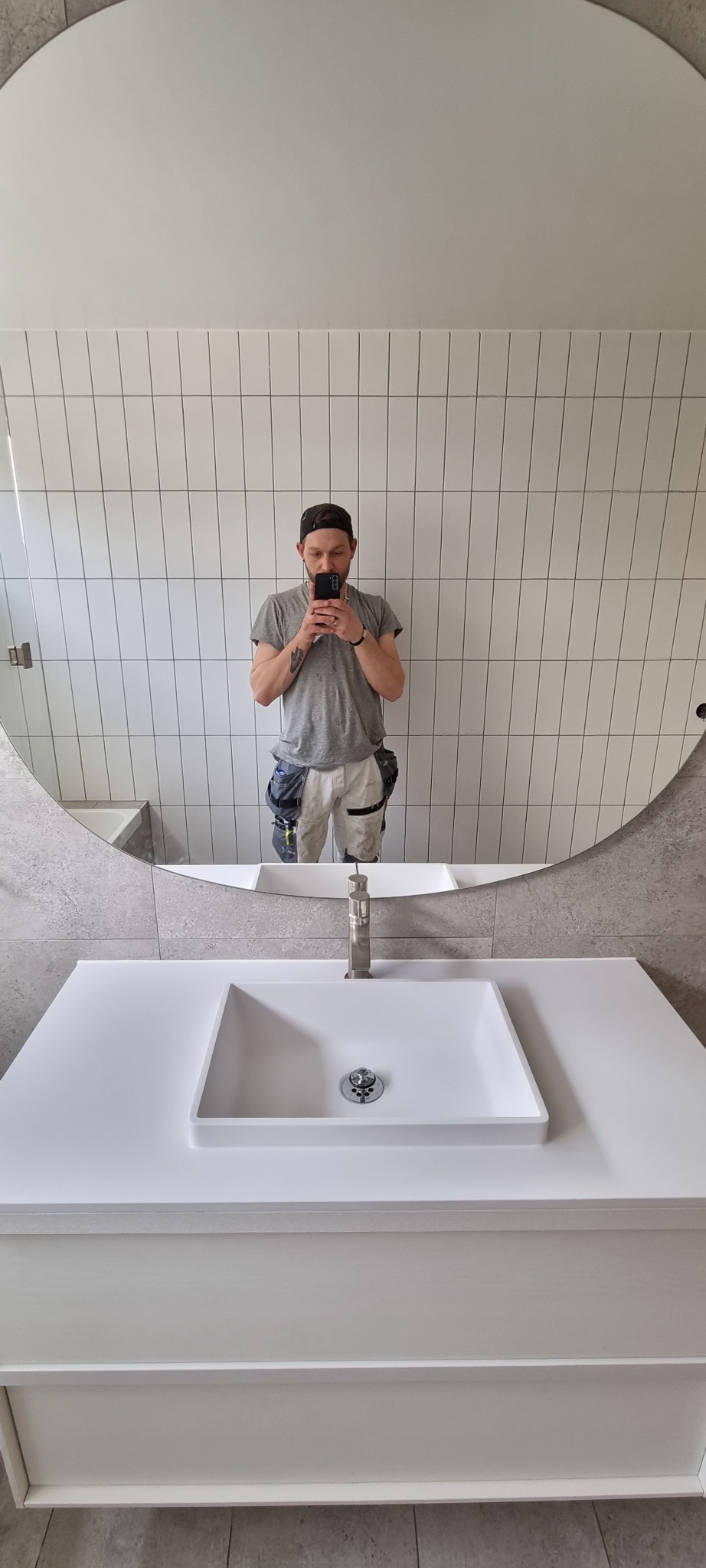 Nowoczesna łazienka z prostokątną umywalką nablatową, okrągłym lustrem i białymi, pionowo ułożonymi płytkami na ścianie. Widoczne odbicie osoby robiącej zdjęcie telefonem.