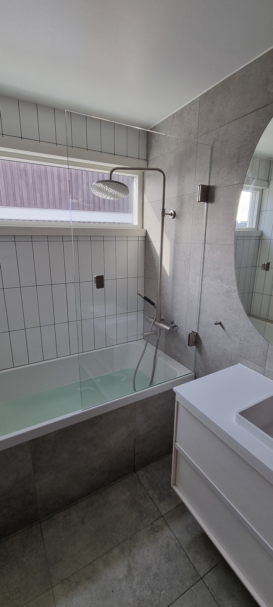 Nowoczesna łazienka z wanną wypełnioną wodą, szklanym parawanem, deszczownicą i okrągłym lustrem, wykończona szarymi płytkami w dwóch odcieniach, z widoczną szafką pod umywalkę w kolorze białym.