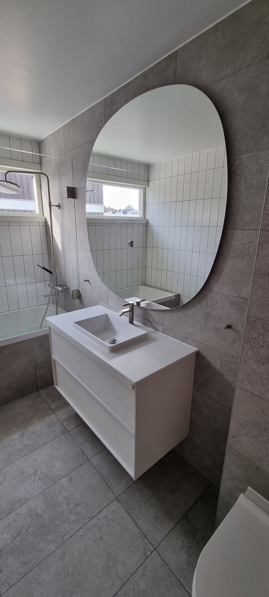 Nowoczesna łazienka z szarymi płytkami, owalnym lustrem i białą szafką pod umywalką z dwoma szufladami, widoczna bateria umywalkowa, w tle wanna i okno.