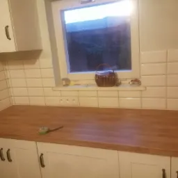 Jasna kuchnia z drewnianym blatem, białymi szafkami i ceramicznymi płytkami na ścianie, widoczny fragment okna z koszykiem i drobiazgami na parapecie oraz miarka na blacie.