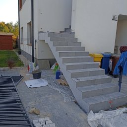 Artur Lech - Zewnętrzne schody z szarych płytek prowadzące do domu, widoczne materiały budowlane i narzędzia, kostka brukowa w trakcie układania, częściowo zakryte folią ochronną.