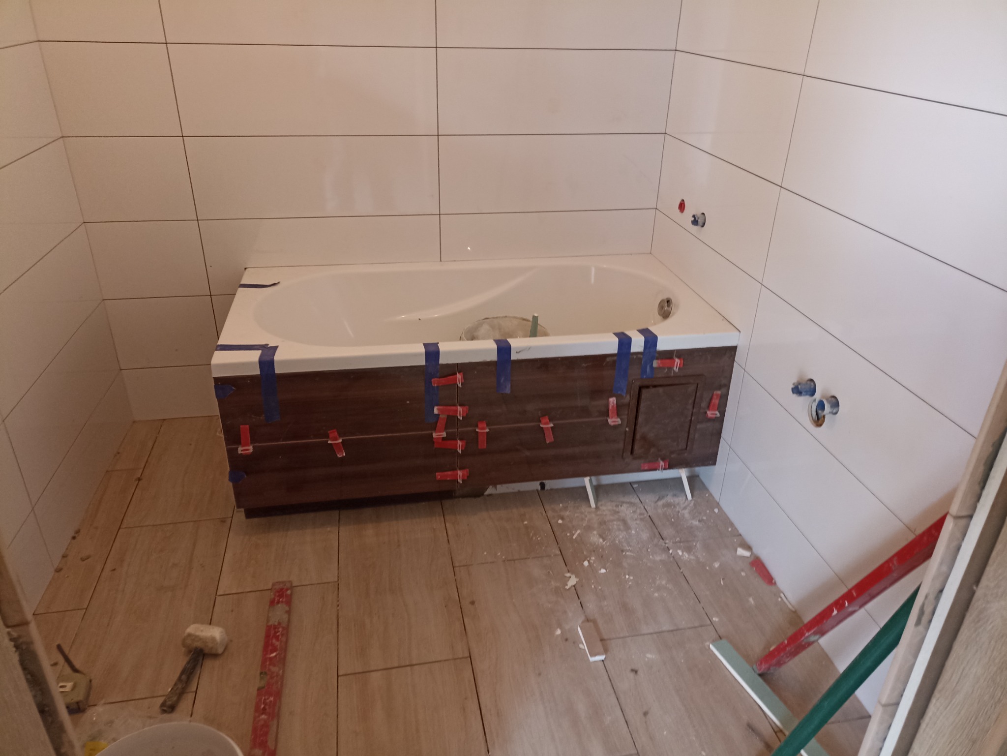 Biała wanna z obudową z płytek imitujących drewno, zabezpieczona niebieską taśmą i czerwonymi krzyżykami dystansowymi, w trakcie układania płytek na ścianach i podłodze. Widoczne przyłącza wodne...