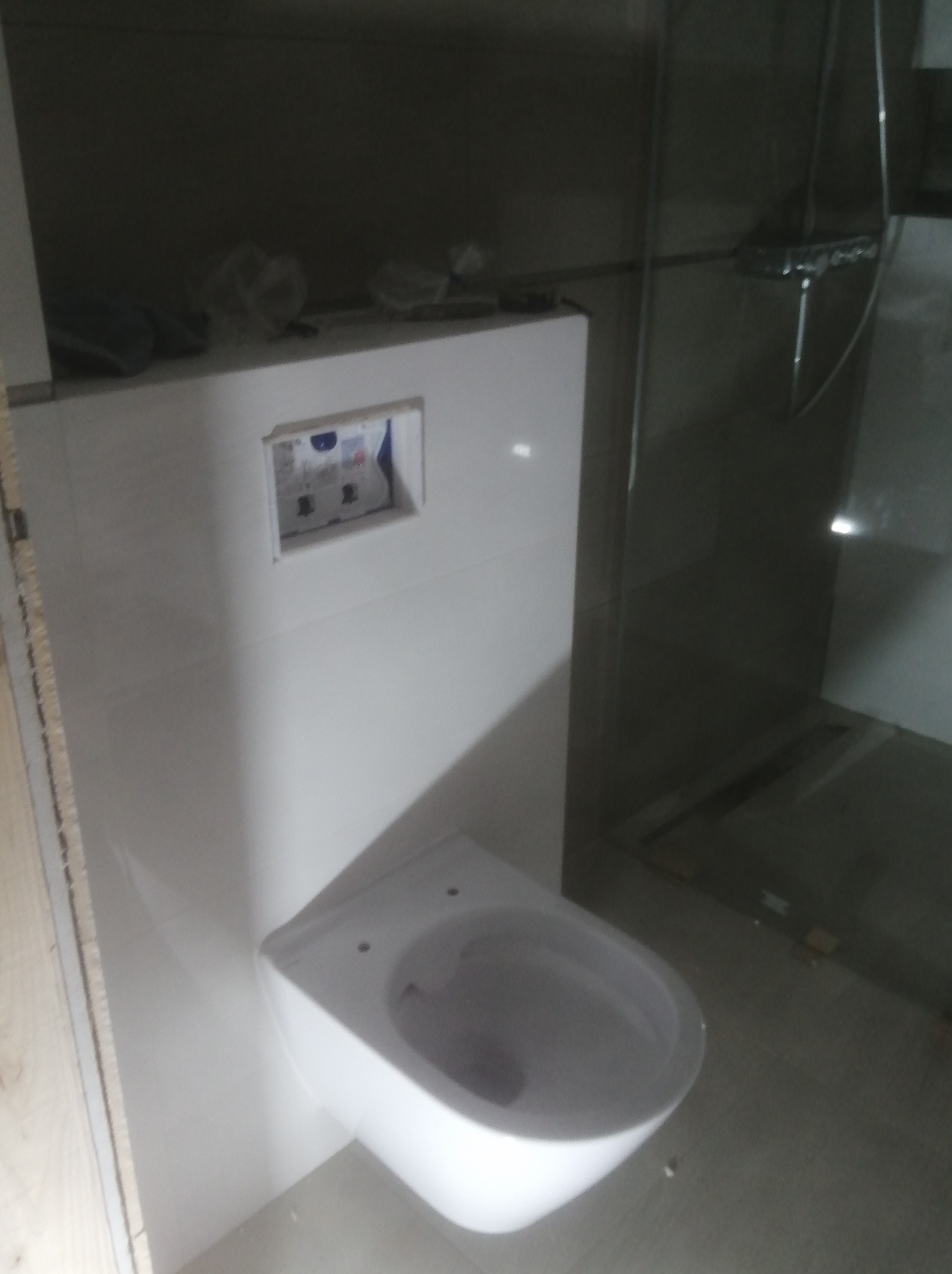 Wisząca miska WC zamontowana na stelażu podtynkowym w trakcie prac wykończeniowych łazienki, widoczne przyłącza i częściowo ułożone płytki na ścianie. W tle widoczny prysznic.