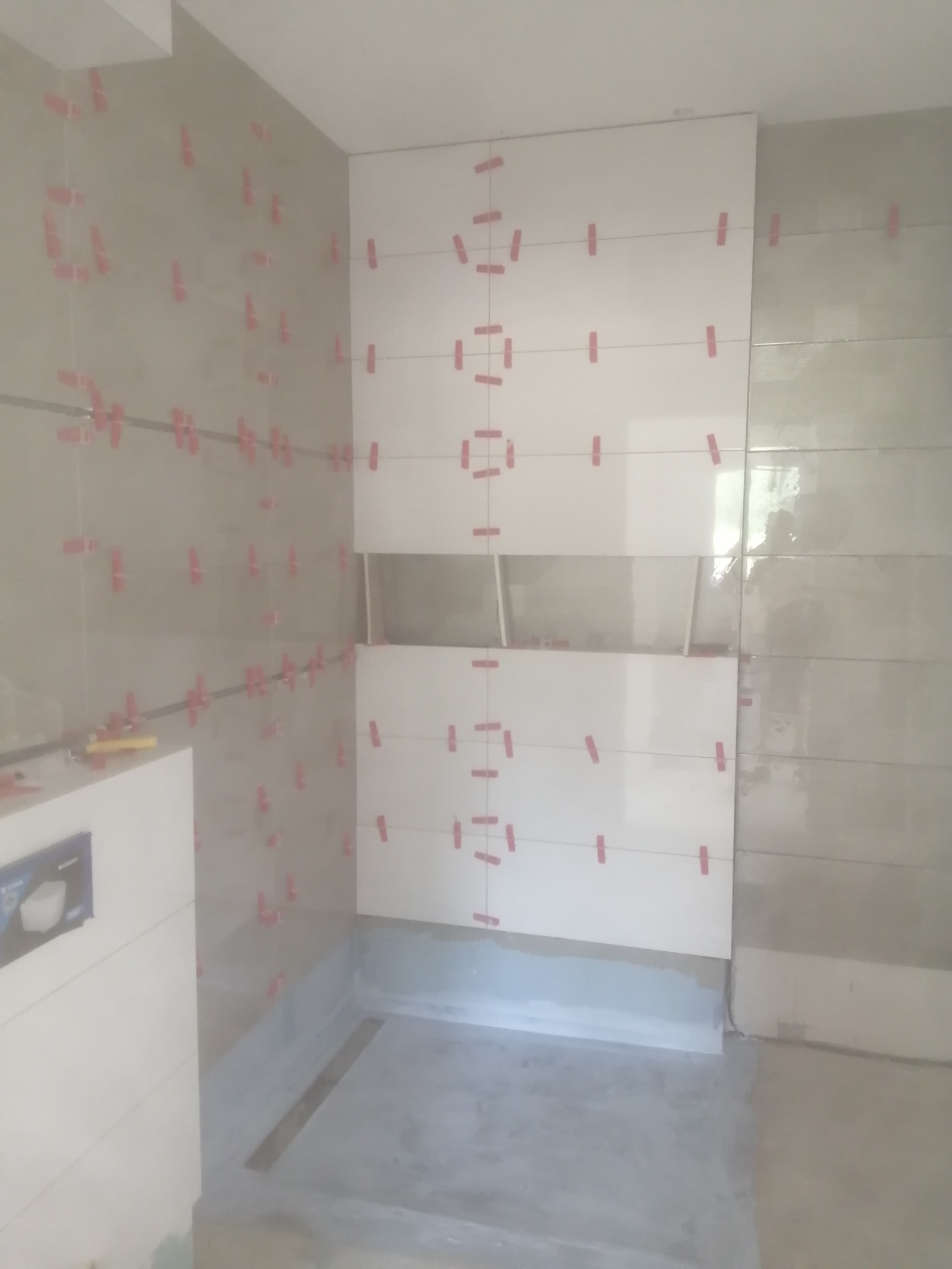 Ściana łazienki w trakcie układania białych płytek, z widocznymi krzyżykami dystansowymi i poziomowaniem laserowym, częściowo wykończona szara ściana w tle.