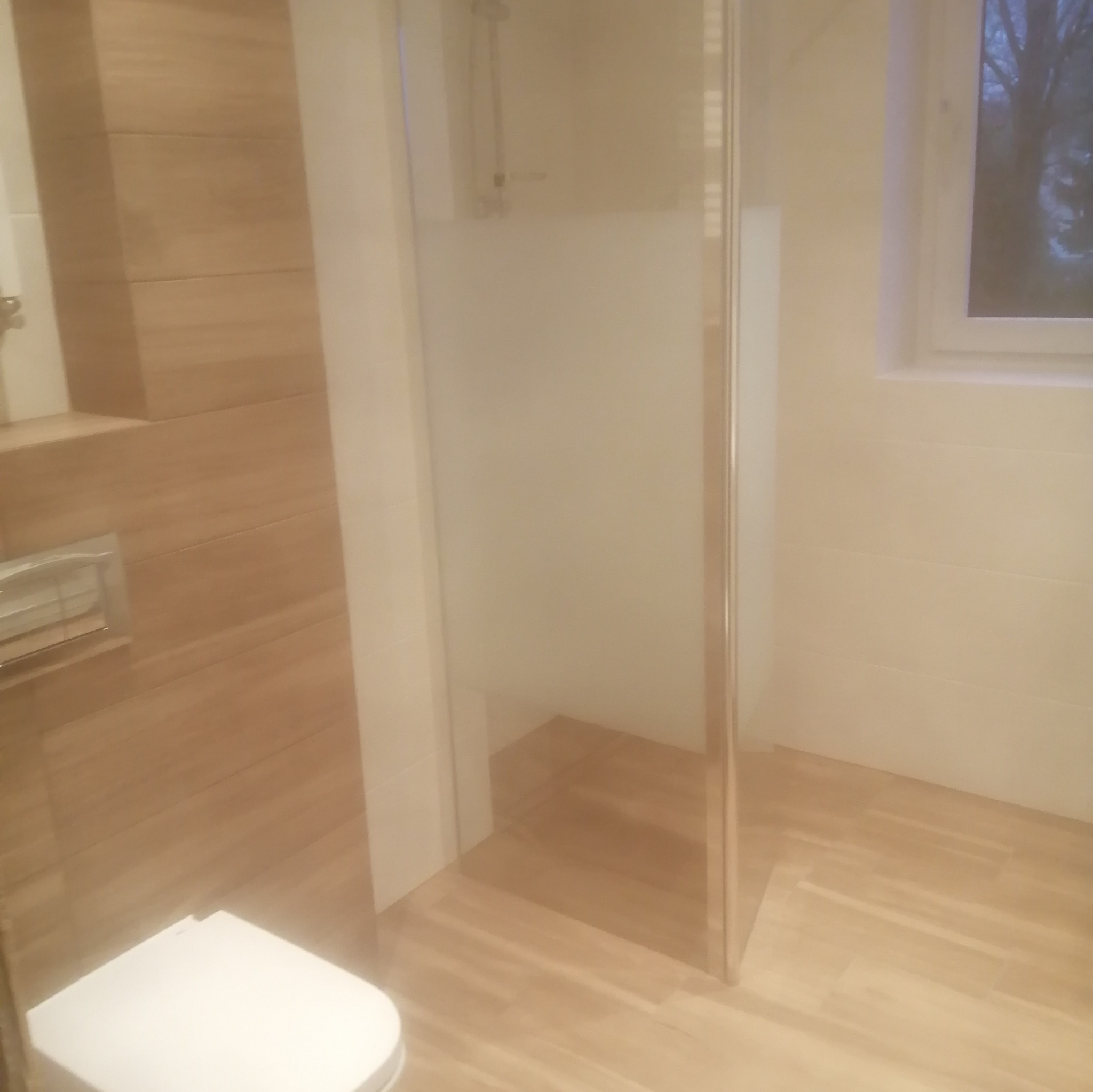 Nowoczesna łazienka z kabiną prysznicową typu walk-in, wykończona płytkami imitującymi drewno na ścianach i podłodze, widoczna toaleta w rogu i okno z widokiem na drzewa.