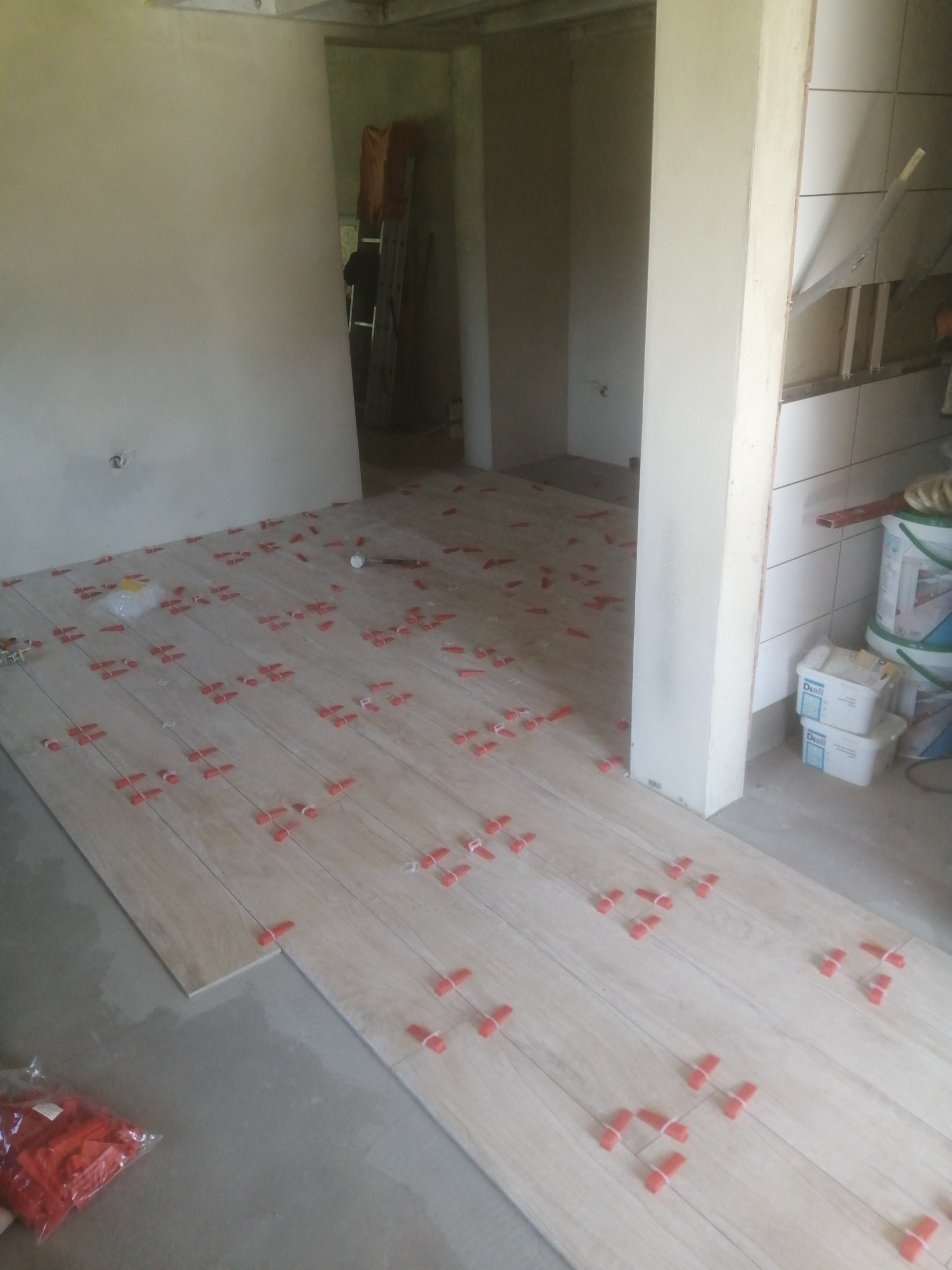 Układanie płytek podłogowych imitujących drewno, z użyciem czerwonych krzyżyków dystansowych, widok na surową ścianę i materiały budowlane w tle.