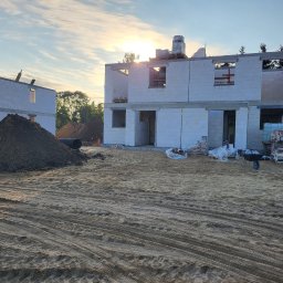 WALER OLEKSANDR KARPIUK - Wschodzące słońce oświetla surową konstrukcję domu z białych bloczków, z widocznymi stertami ziemi i materiałów budowlanych na piaszczystym terenie budowy.