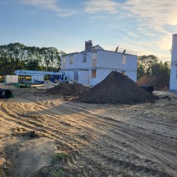 WALER OLEKSANDR KARPIUK - Budowa domu z białego bloczka: ściany w stanie surowym, widoczny dźwig, hałdy ziemi i rury kanalizacyjne. W tle drzewa i błękitne niebo.