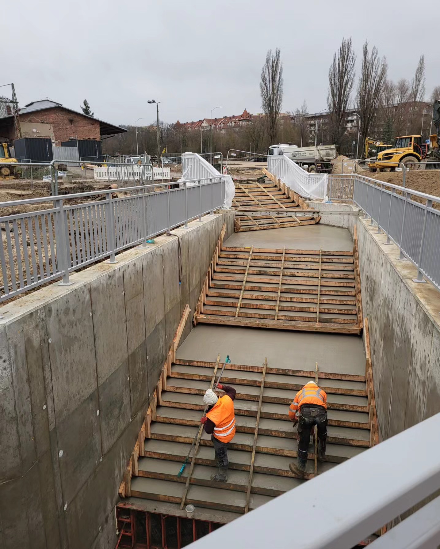 Betonowanie schodów w przejściu podziemnym. Dwóch robotników w odblaskowych kamizelkach pracuje na drewnianym szalunku. Widoczne maszyny budowlane w tle.