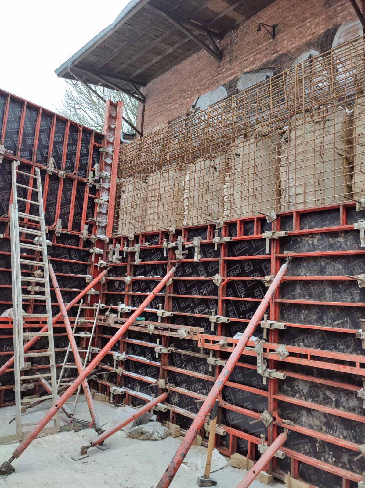 Szczegółowe ujęcie szalunków i zbrojenia na budowie, z widoczną drabiną i fragmentem ceglanego budynku w tle. Elementy konstrukcyjne PERI.