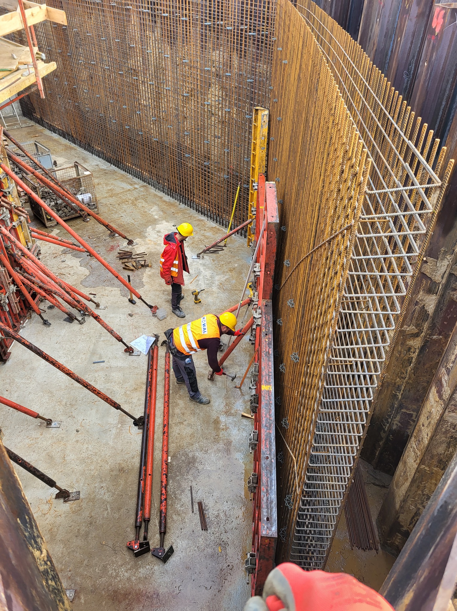 Praca zbrojarzy przy formowaniu szalunku i zbrojenia pionowej ściany wykopu. Widoczne stalowe pręty zbrojeniowe, podpory szalunkowe i dwóch pracowników w kaskach.