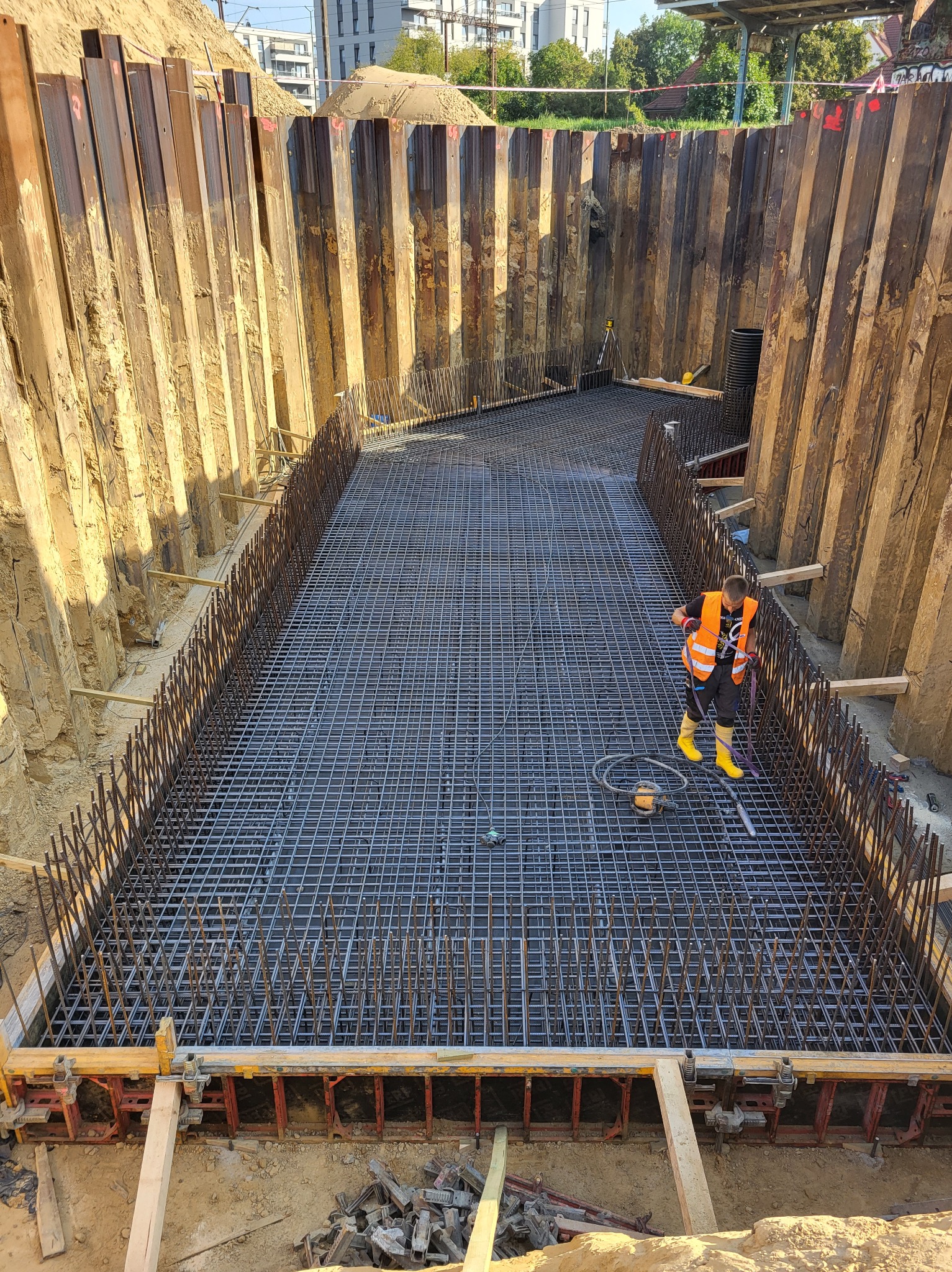 Zbrojenie fundamentu w wykopie zabezpieczonym ściankami Larsena. Pracownik w żółtych butach i kamizelce. Widoczne narzędzia i elementy szalunku.