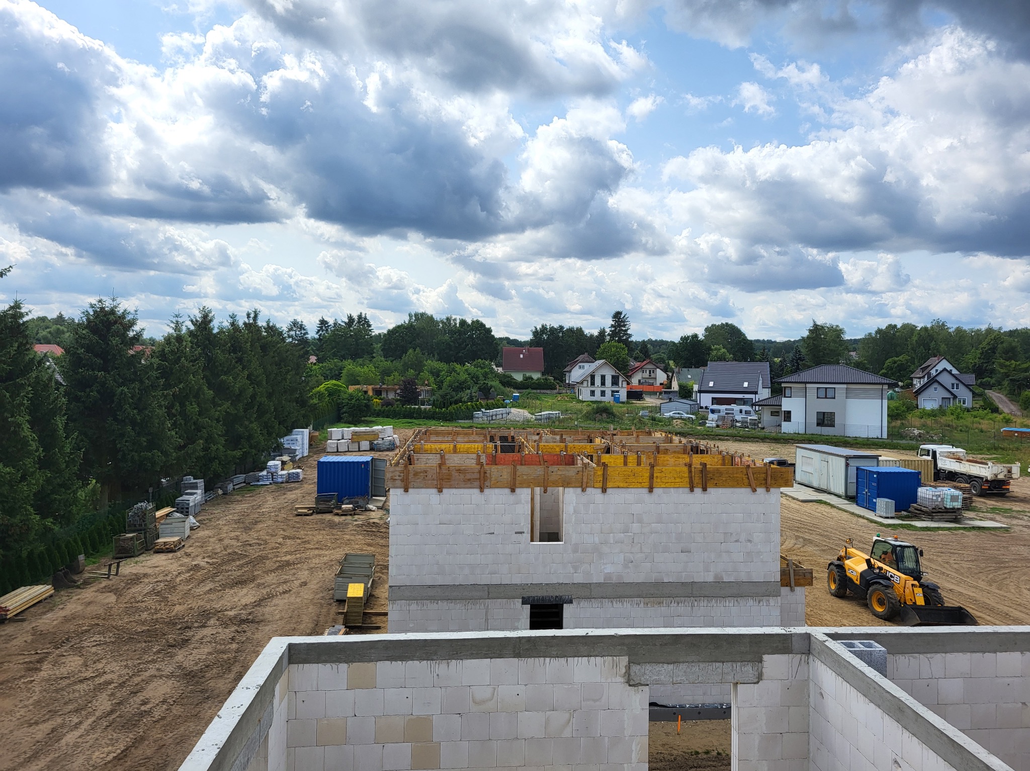 Wznoszenie ścian z betonu komórkowego na budowie domu jednorodzinnego. Widoczny szalunek i maszyna budowlana. W tle zabudowa jednorodzinna i zieleń.