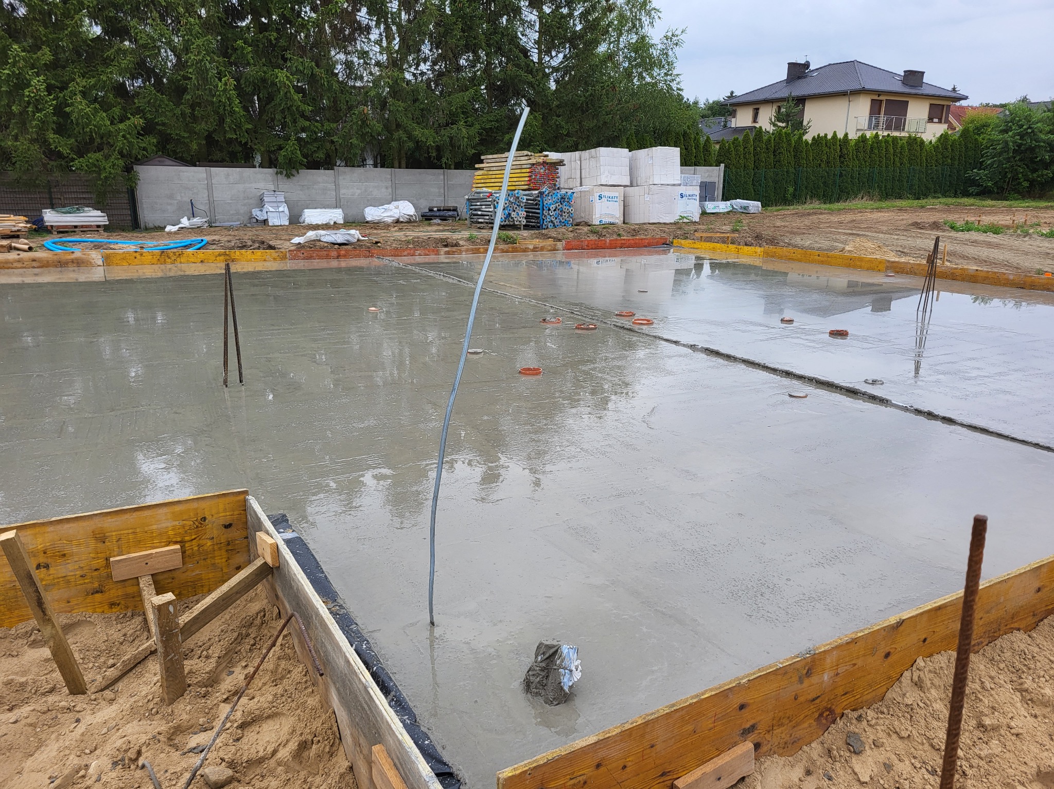 Zalany fundament pod budowę domu jednorodzinnego. Widoczne szalunki, wystające pręty zbrojeniowe i elementy instalacji. W tle materiały budowlane i dom.