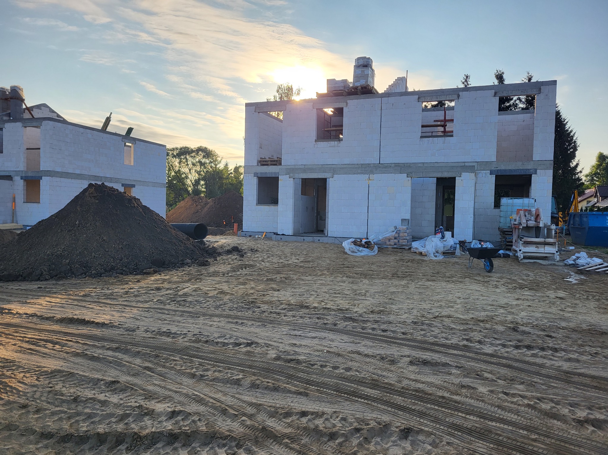 Wschodzące słońce oświetla surową konstrukcję domu z białych bloczków, z widocznymi stertami ziemi i materiałów budowlanych na piaszczystym terenie budowy.