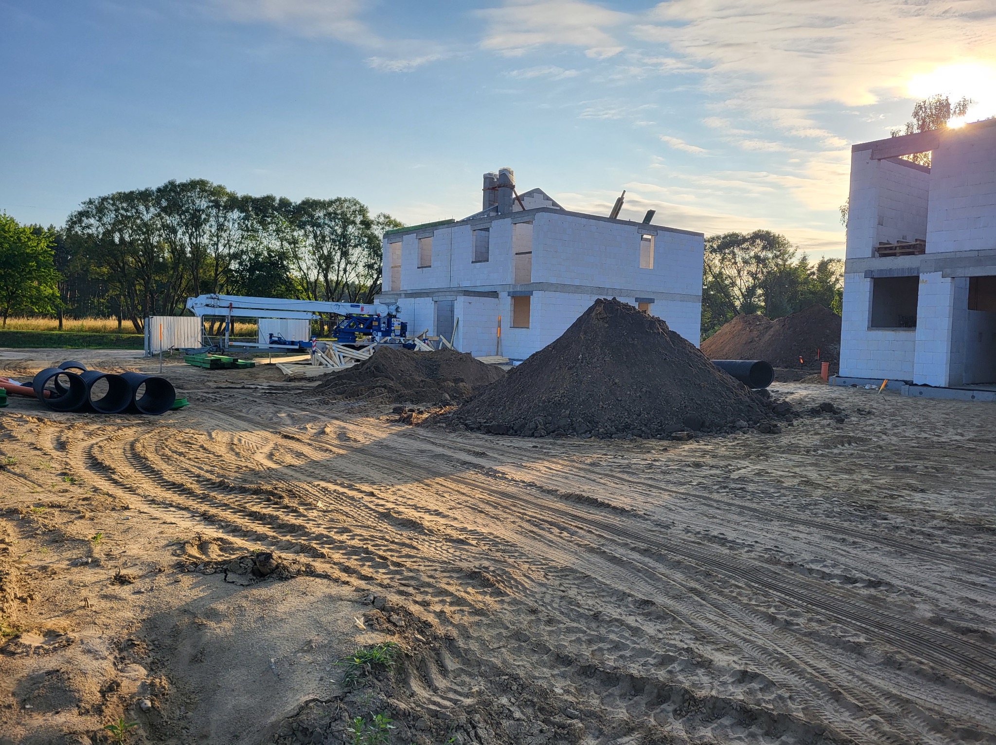 Budowa domu z białego bloczka: ściany w stanie surowym, widoczny dźwig, hałdy ziemi i rury kanalizacyjne. W tle drzewa i błękitne niebo.