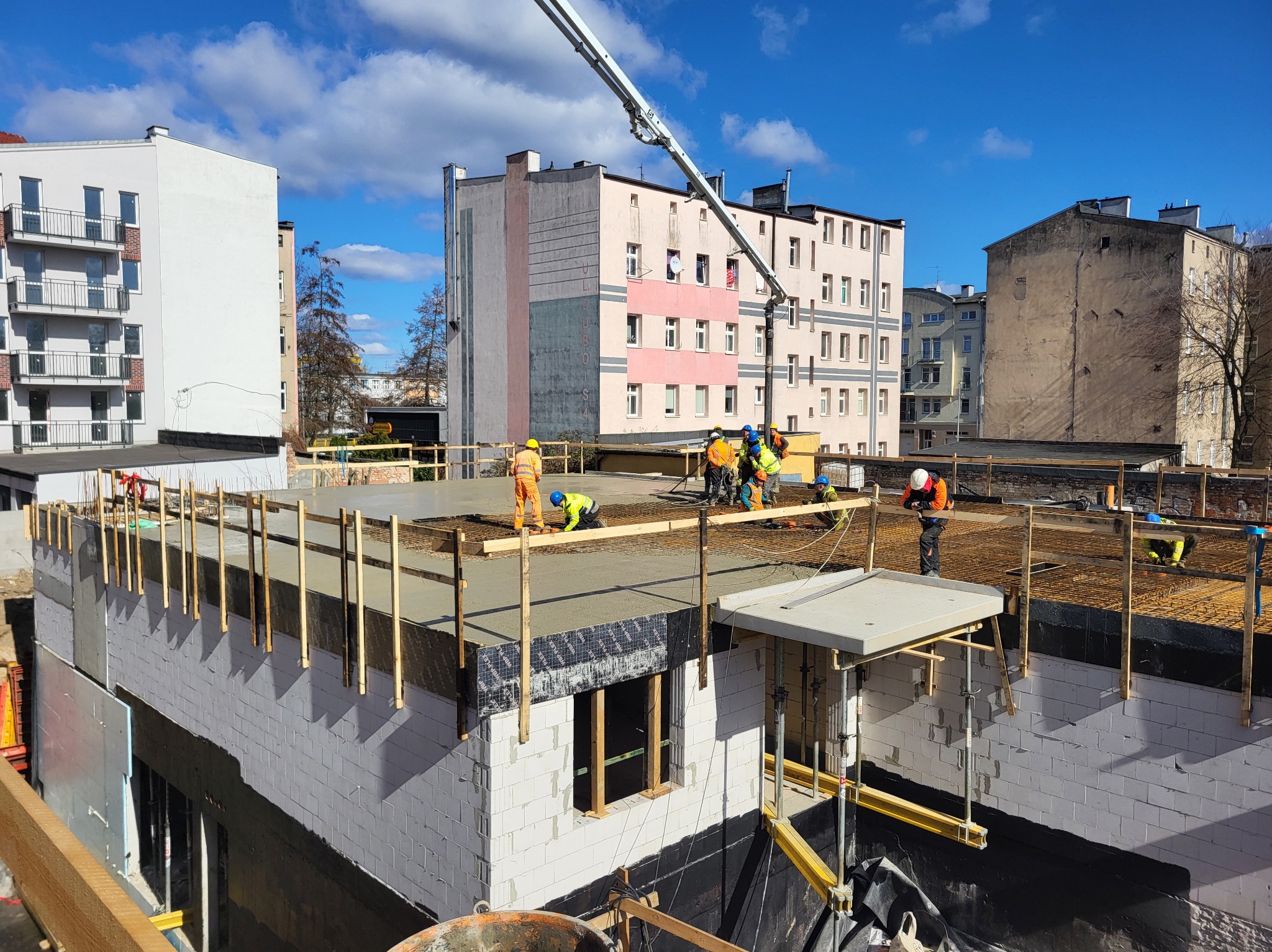 Wylewanie betonu na strop budynku z białego bloczka. Widoczni pracownicy w kaskach i kamizelkach, pompa do betonu, zbrojenie. Słoneczny dzień, budynki w tle.