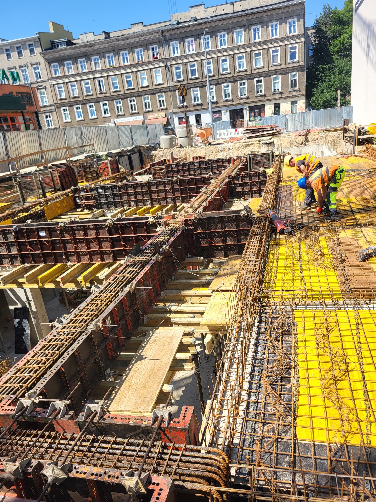 Zbrojenie stropu na budowie z widocznymi deskowaniami i pracującymi zbrojarzami w odblaskowych kamizelkach. W tle budynek miejski i błękitne niebo.