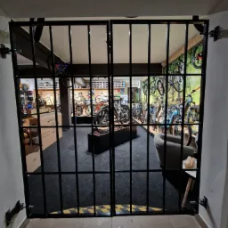 Zamykana, metalowa krata w kolorze czarnym, zabezpieczająca wejście do sklepu z rowerami, widoczne liczne modele rowerów wewnątrz.