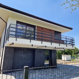 MetalFam Jakub Konfederak - Nowoczesny dom z balkonem zabezpieczonym czarną balustradą poziomą, wykończony grafitową okładziną elewacyjną, widziany z poziomu gruntu przez ogrodzenie.