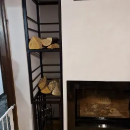 Metalowy regał na drewno, czarny, z dwoma półkami wypełnionymi drewnem kominkowym, stojący obok kominka z czarną obudową, widoczne narzędzia kominkowe w dolnej części regału.