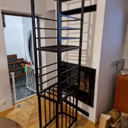 Metalowa konstrukcja regału na kółkach, z czarnych profili, w surowym wnętrzu z parkietem i kominkiem.