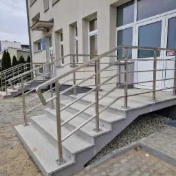 Zewnętrzne schody wejściowe do budynku z granitowymi stopniami i balustradą ze stali nierdzewnej o poziomych wypełnieniach, widoczny fragment elewacji, okna i drzwi wejściowe.