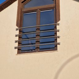 MetalFam Jakub Konfederak - Okno na poddaszu z brązową ramą i zabezpieczeniem w postaci poziomych, metalowych prętów mocowanych do ramy za pomocą okrągłych uchwytów.