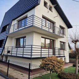 MetalFam Jakub Konfederak - Nowoczesny dom z balkonami zabezpieczonymi czarnymi, prostymi balustradami, widok z dołu na elewację z kontrastującym, ciemnym dachem i jasnymi ścianami.