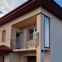 Nowoczesny dom z beżową elewacją, balkonem z czarną metalową balustradą i wysokim oknem narożnym.