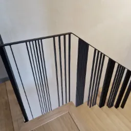 Nowoczesne schody z drewnianymi stopniami i czarną, minimalistyczną balustradą z pionowych prętów, widziane z góry pod kątem.